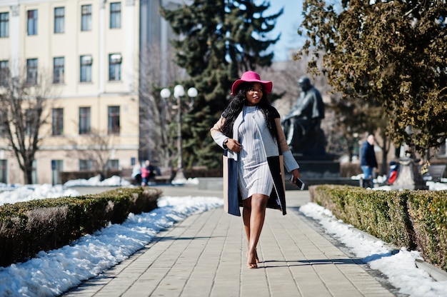 Mujer afroamericana en sombrero y abrigo con teléfono caminando en las calles.