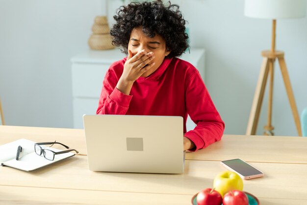 Mujer afroamericana soñolienta sentada en la mesa con una computadora portátil y bostezando cansada chica con exceso de trabajo perezosa para