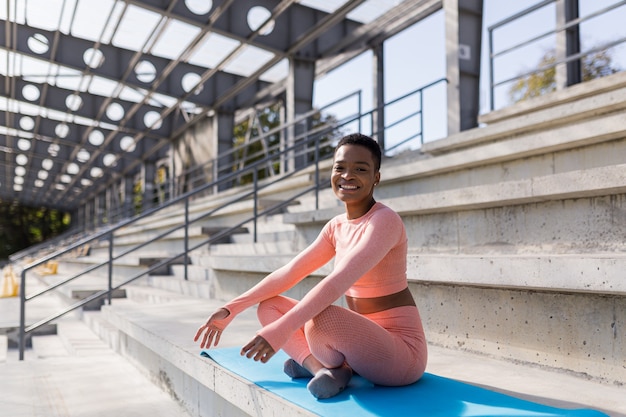 Mujer afroamericana sonriendo y mirando a la cámara, sentada en posición de loto en la estera de fitness, confiada y alegre después del yoga