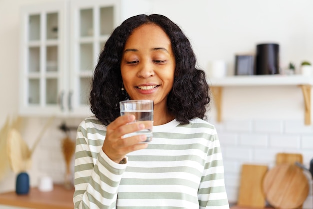 Una mujer afroamericana sonriente disfrutando de un vaso de agua clara mientras está de pie en la cocina