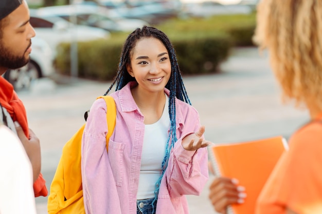 Mujer afroamericana sonriente estudiante inteligente sosteniendo mochila amarilla comunicación con amigos