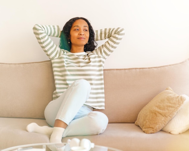 Foto una mujer afroamericana sonriente poniendo las manos detrás de la cabeza mientras está sentada en el sofá en la sala de estar