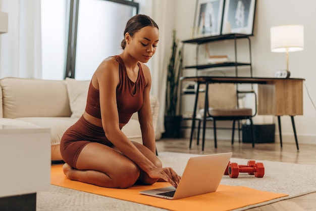 Una mujer afroamericana sonriente que va a practicar yoga usando una computadora portátil en la sala de estar de su casa