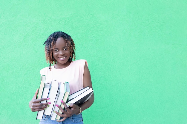 Una mujer afroamericana sonriente sostiene una pila de libros en un espacio de copia de estudiante de fondo verde