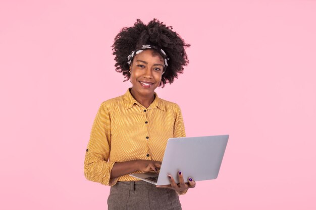 Mujer afroamericana sosteniendo una computadora portátil sonriendo y mirando a la cámara Mujer joven y hermosa sintiéndose feliz de pie aislada sobre un fondo rosa