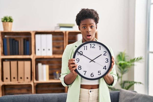 Mujer afroamericana sosteniendo un gran reloj en la cara de shock, luciendo escéptica y sarcástica, sorprendida con la boca abierta