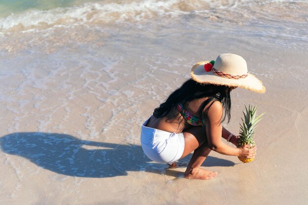 Mujer afroamericana sostiene una piña con el mar de fondo.