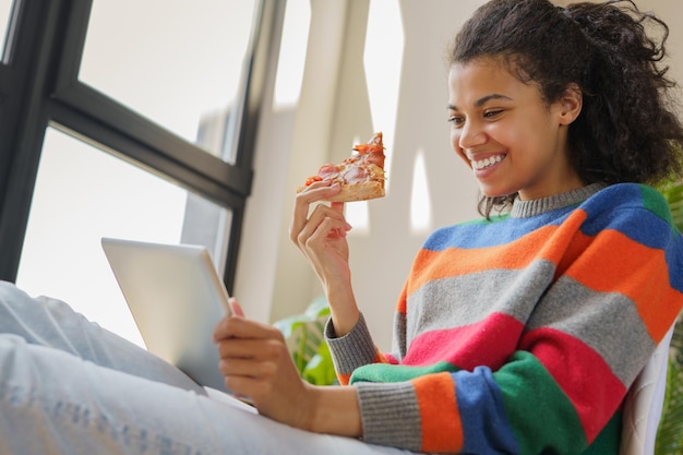 Mujer afroamericana con tableta digital viendo películas en línea comiendo pizza relajándose en casa