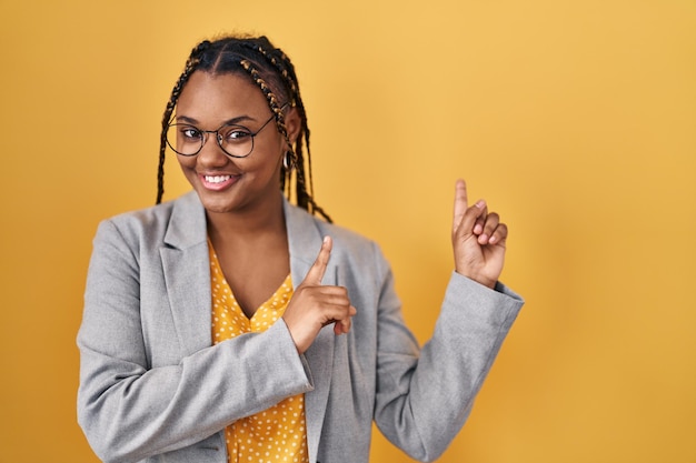 mujer afroamericana con trenzas de pie sobre fondo amarillo sonriendo y mirando a la cámara apuntando con dos manos y dedos hacia el lado