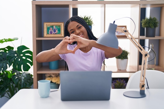 Mujer afroamericana con trenzas usando una laptop en casa sonriendo enamorada mostrando el símbolo del corazón y la forma con las manos. concepto romántico.