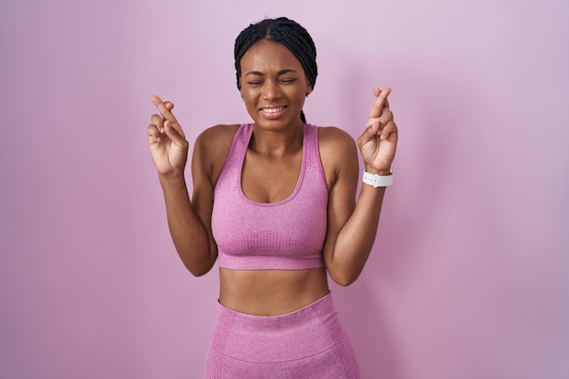 Mujer afroamericana con trenzas usando ropa deportiva sobre fondo rosa haciendo gestos con los dedos cruzados sonriendo con esperanza y ojos cerrados suerte y concepto supersticioso