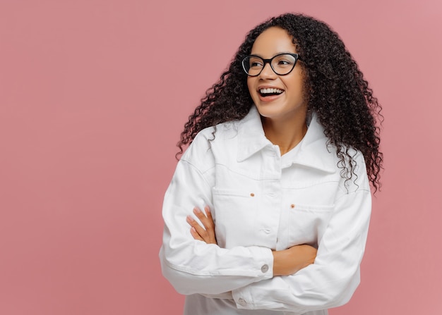 La mujer afroamericana usa una chaqueta blanca, mantiene el brazo doblado, mira a un lado, tiene una sonrisa encantadora