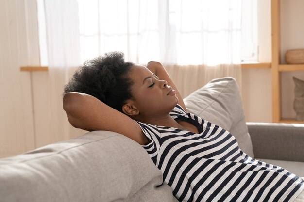 Mujer afroamericana usar camiseta despojada durmiendo en el sofá con el brazo debajo de la cabeza en casa. Descanso.
