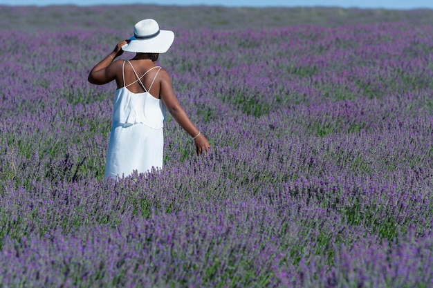 Mujer afroamericana con vestido blanco y sombrero paseando entre las filas de lavanda en flor