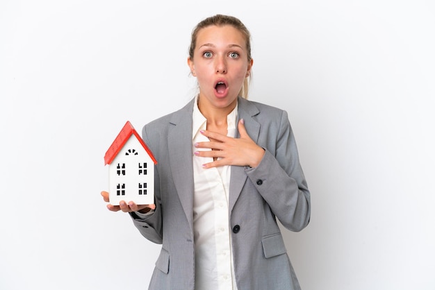 Mujer agente de bienes raíces sosteniendo una casa de juguete aislada de fondo blanco sorprendida y conmocionada mientras miraba a la derecha