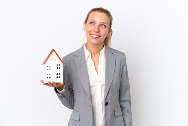 Foto mujer agente inmobiliaria sosteniendo una casa de juguete aislada de fondo blanco pensando en una idea mientras mira hacia arriba