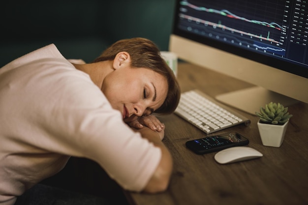 Mujer agotada de mediana edad durmiendo en su escritorio en casa durante una noche trabajando y comerciando con criptomonedas.