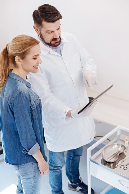 Mujer agradable atractiva alegre de pie cerca de su médico y mirando la pantalla de la tableta mientras se sale de su cirugía estética