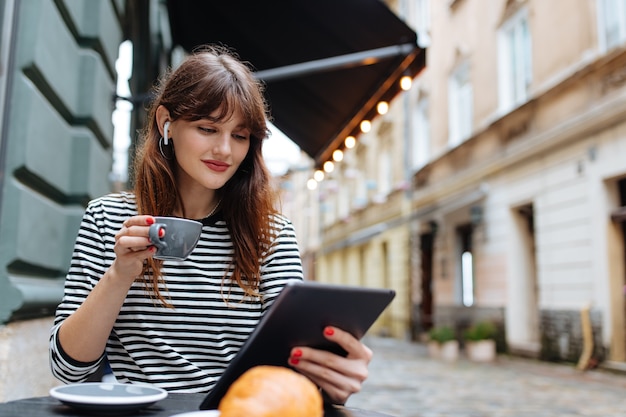 Mujer agradable con cabello castaño mirando la pantalla de la tableta mientras se relaja en el acogedor café