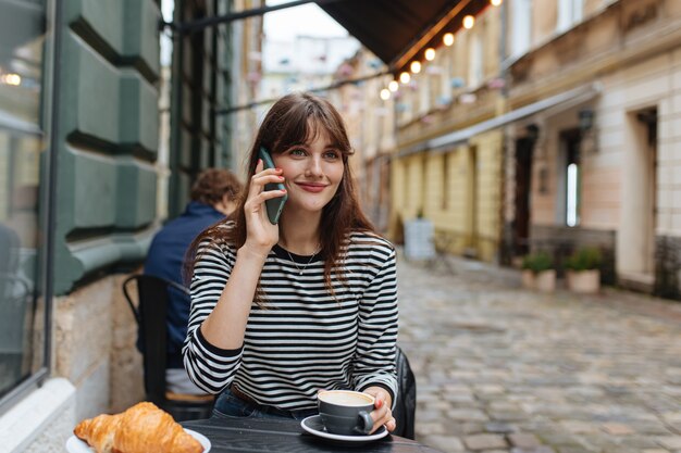 Mujer agradable en suéter a rayas que tiene una conversación móvil mientras toma un café en la terraza del café