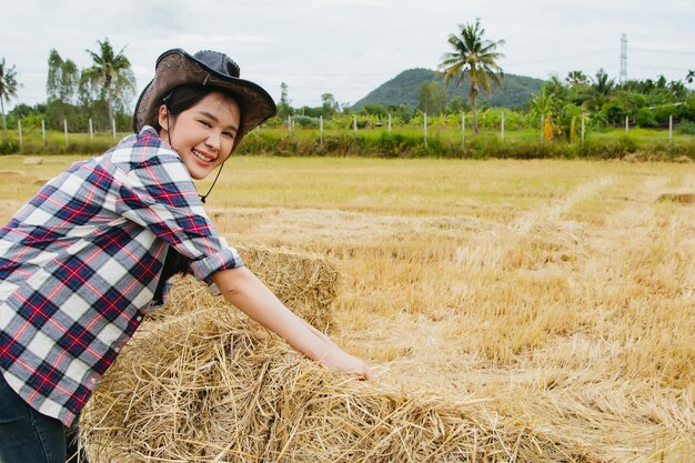 Mujer agricultora asiática sana con sombrero de trabajo al aire libre recogiendo paja en el campo de arroz disfrutando de la cosecha.