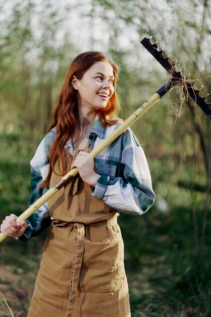 Mujer agricultora bellamente sonriente con ropa de trabajo y delantal trabajando al aire libre en la naturaleza y sosteniendo un rastrillo para recoger hierba