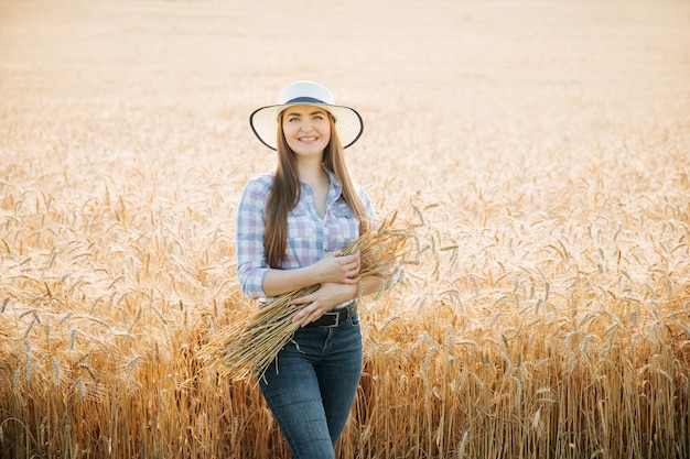 Mujer agricultora con sombrero en la cabeza se encuentra en campo con trigo amarillo y sostiene espigas de trigo y ...