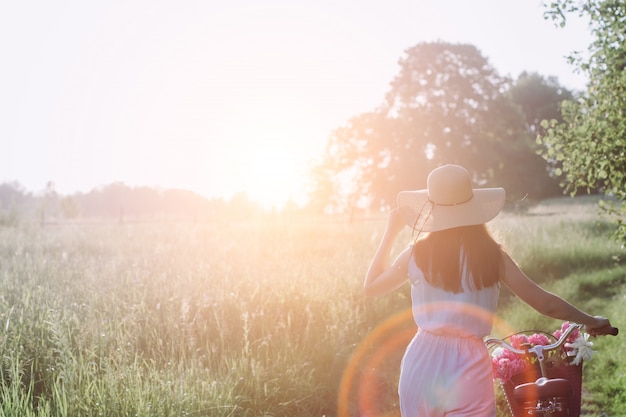 Mujer al aire libre con bicicleta vintage y una cesta de flores y disfrutando del atardecer contra