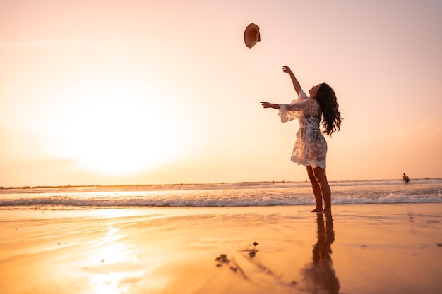 Una mujer al atardecer con un vestido blanco en el mar en verano tirando el sombrero de vacaciones