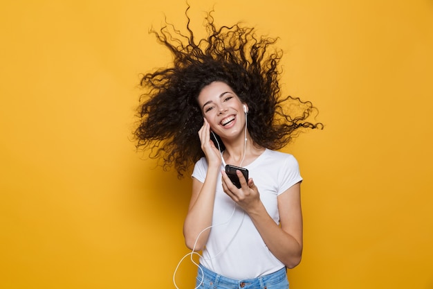 Mujer alegre de 20 años con cabello rizado sacudiendo el teléfono inteligente y escuchando música a través de auriculares aislados en amarillo