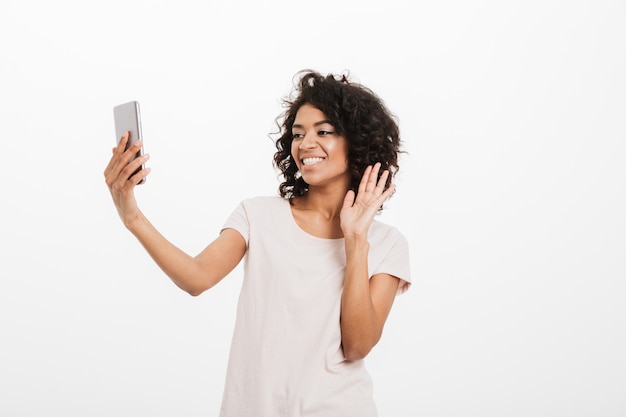 Mujer alegre de 20 años con el pelo rizado con camiseta básica con teléfono móvil y sonriendo mientras toma selfie, aislado sobre pared blanca