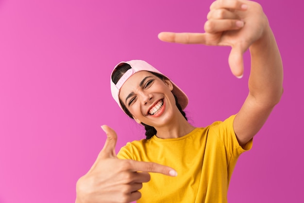 Mujer alegre alegre vistiendo gorra riendo y mostrando el marco vertical con los dedos aislados en la pared rosa