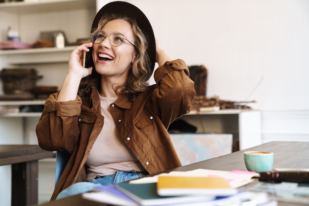 Mujer alegre en anteojos y sombrero hablando por teléfono celular mientras estudia con libros de ejercicios en casa