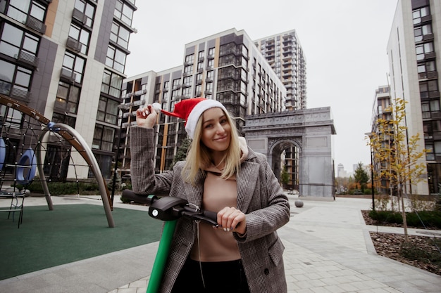 Mujer alegre arreglando su sombrero de santa y conduciendo scooter eléctrico. Bloques de apartamentos en el fondo. Mujer feliz se compró un scooter eléctrico en honor a las vacaciones de Navidad.