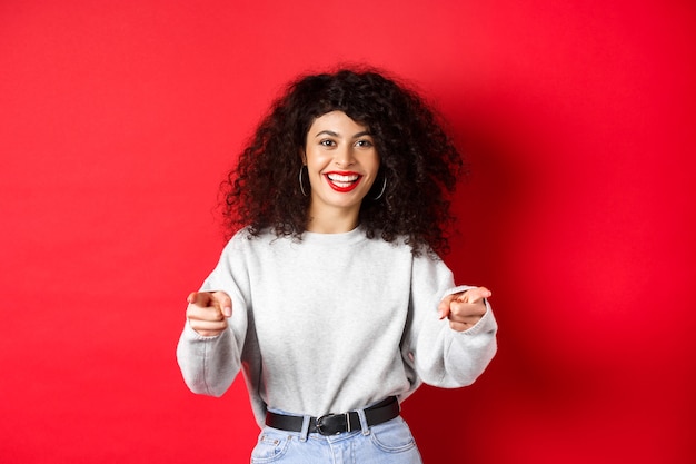 Mujer alegre con cabello rizado invitándote, reclutando novatos, señalando con el dedo a la cámara y sonriendo, alabando el buen trabajo, de pie sobre fondo rojo.