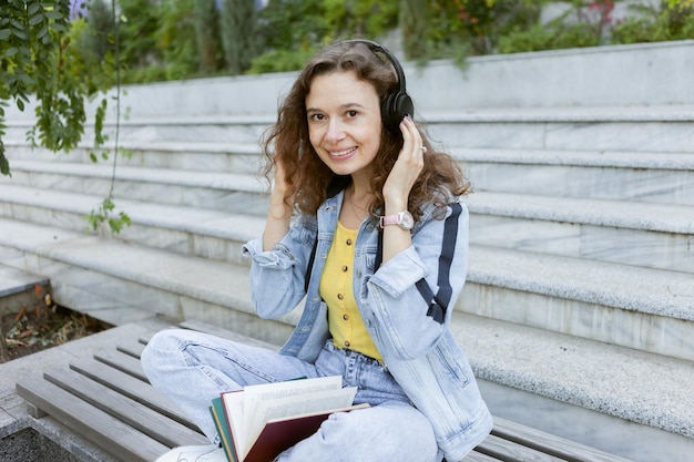 Mujer alegre de cabello rizado de mediana edad con ropa de mezclilla escuchando música y leyendo un libro mientras se sienta en un banco al fondo de las escaleras al aire libre