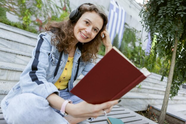 Mujer alegre de cabello rizado de mediana edad con ropa de mezclilla escuchando música y leyendo un libro mientras se sienta en un banco al fondo de las escaleras al aire libre