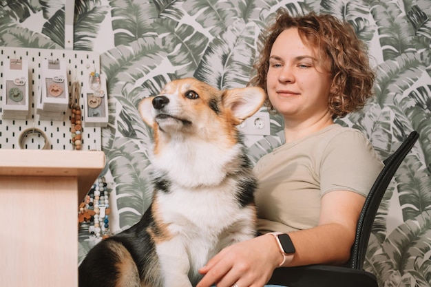 Mujer alegre de cabello rizado propietaria con perro corgi sentado en una silla cerca de una laptop en el lugar de trabajo adentro Tienda de accesorios para bebés