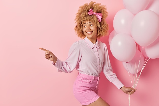 Mujer alegre con cabello rizado usa blusa y pantalones cortos sostiene un montón de puntos de globos inflados a distancia se prepara para la fiesta aislada sobre el espacio vacío de fondo rosa para su promoción