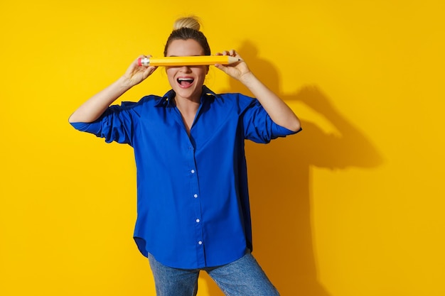 Mujer alegre con camisa azul sosteniendo un lápiz gigante sobre fondo amarillo