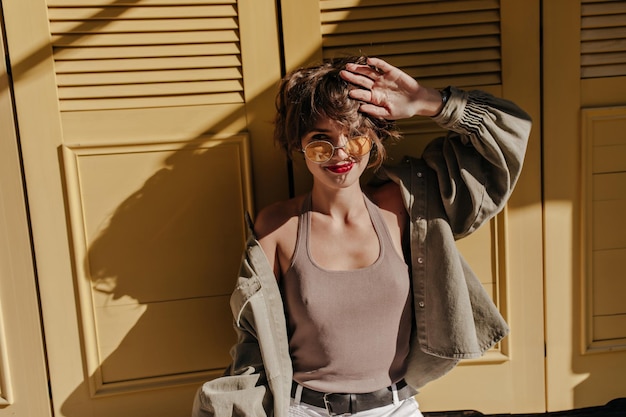 Mujer alegre en camiseta y chaqueta de mezclilla posando sobre fondo brillante Chica rizada en gafas de sol redondas con labios rojos sobre fondo amarillo