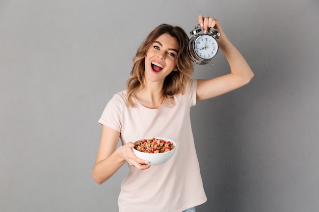 Mujer alegre en camiseta con comida sana en plato y despertador mientras sobre gris
