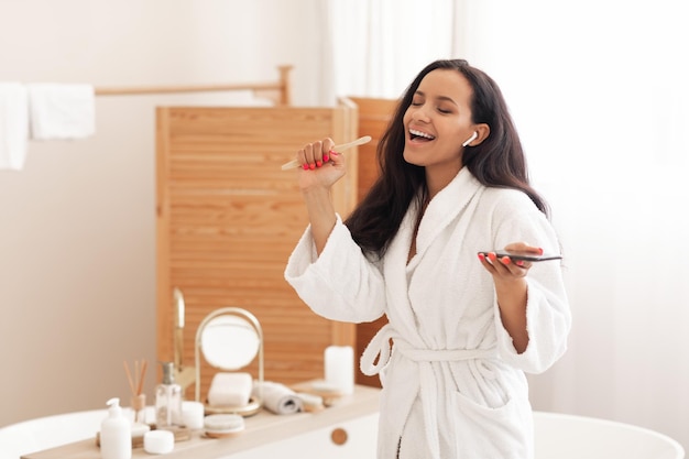 Mujer alegre cantando sosteniendo cepillo de dientes de madera y teléfono en el baño.