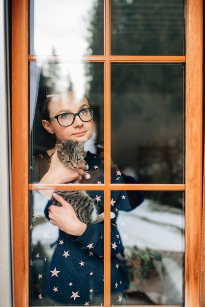 Mujer alegre con cara bonita tiene un gato encantador en sus manos y mira por la ventana