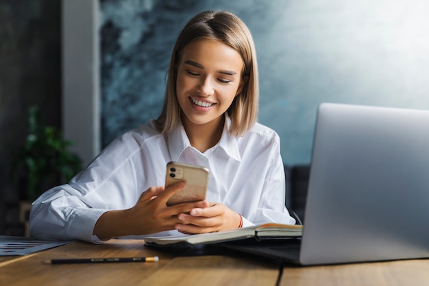 Mujer alegre con descanso en el lugar de trabajo, comunicándose con amigos en aplicaciones de mensajería