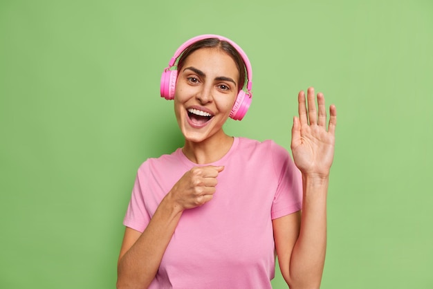 La mujer alegre y despreocupada canta su canción favorita mantiene la mano cerca de la boca como si el micrófono escuchara la pista de audio en los auriculares vestidos casualmente posa contra la pared verde vivo