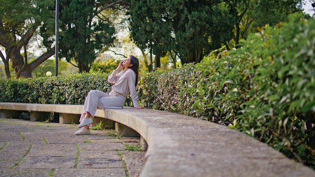 Foto mujer alegre disfrutando de una conversación telefónica sentada en un banco del parque en el follaje verde