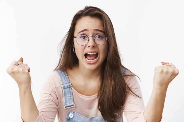 Mujer alegre, emocionada y llena de energía que grita de alegría y apoyo apretando los puños emocionada mientras grita que quiere que el equipo favorito gane siendo un verdadero fanático de pie sobre una pared gris