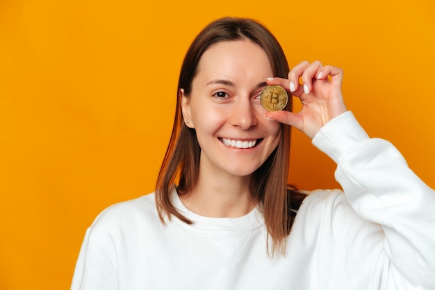 Una mujer alegre esconde un ojo detrás de una moneda dorada de bitcoin mientras sonríe