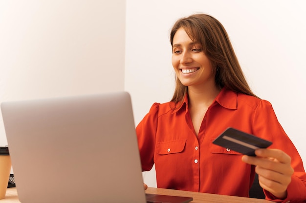 Mujer alegre está comprando en línea mientras escribe en la computadora portátil y sostiene la tarjeta